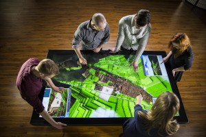 Five users gather around a large multitouch table displaying a 3D model of Cusco
